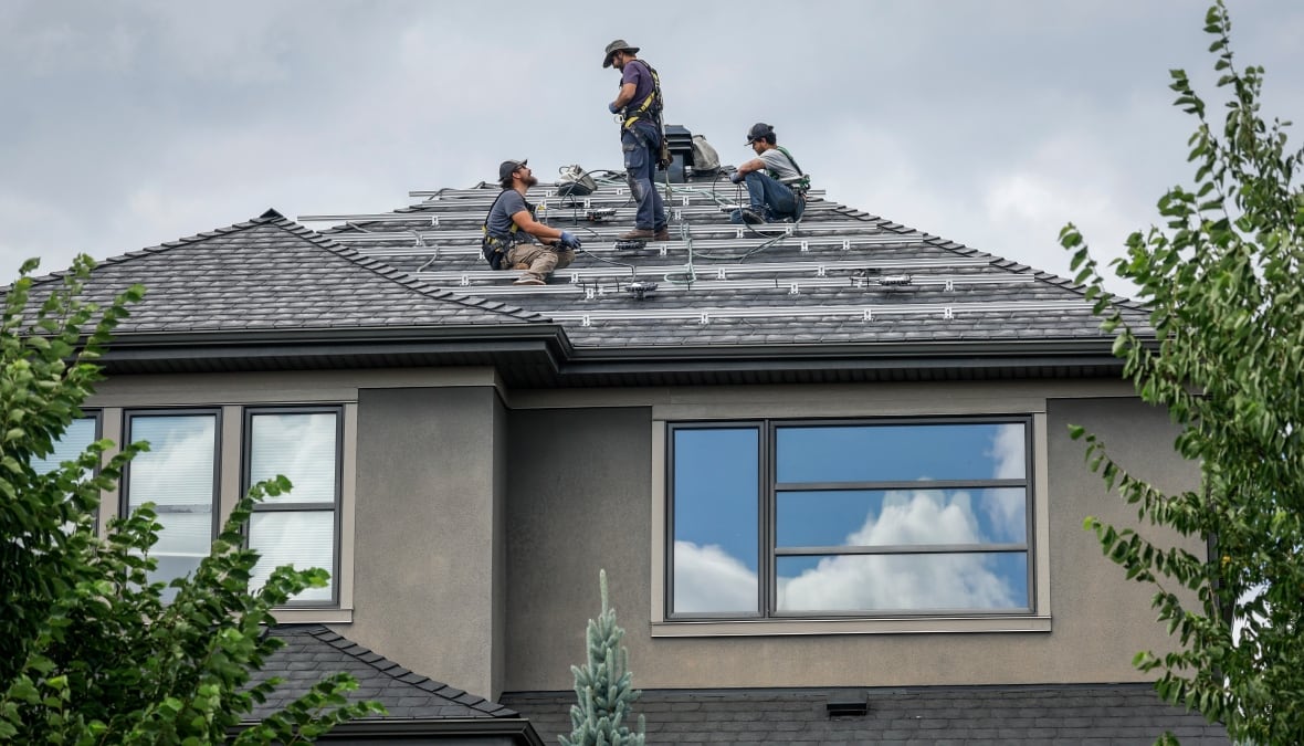Three workers installing metal rails across a shingled rooftop of a house. 