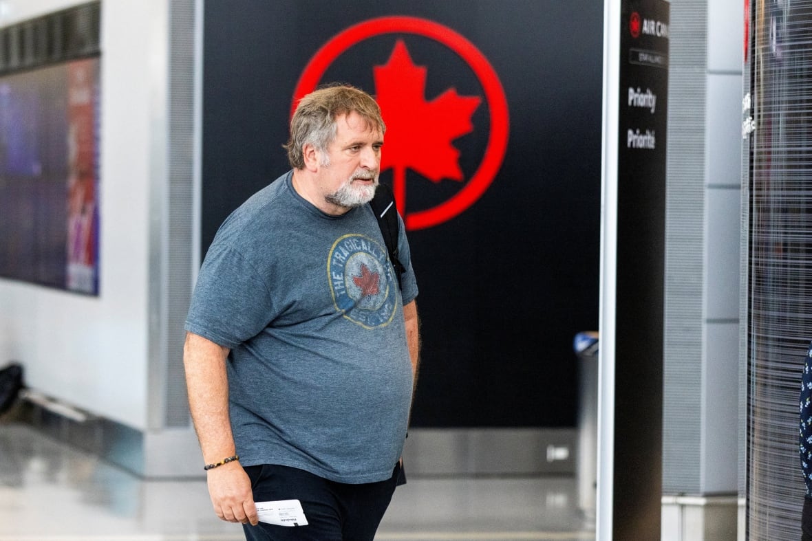 A man in a t-shirt walks in an airport, in front of an Air Canada logo.