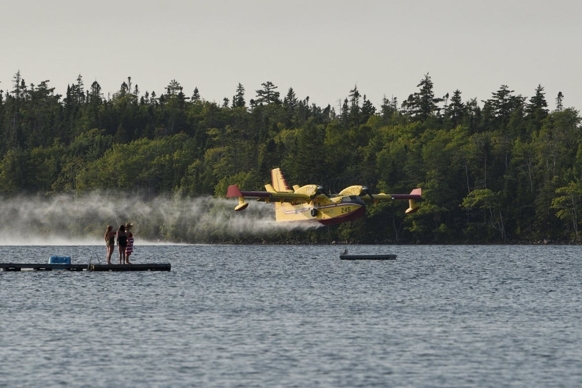 A yellow plane flying very close to a lake as three people watch from a dock.