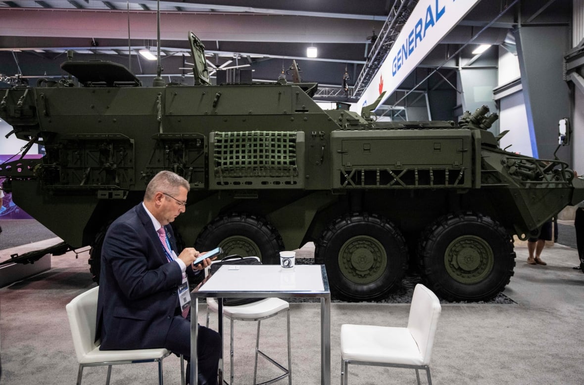 A large military vehicle is seen in a showroom. A man sits at a table in front of it.