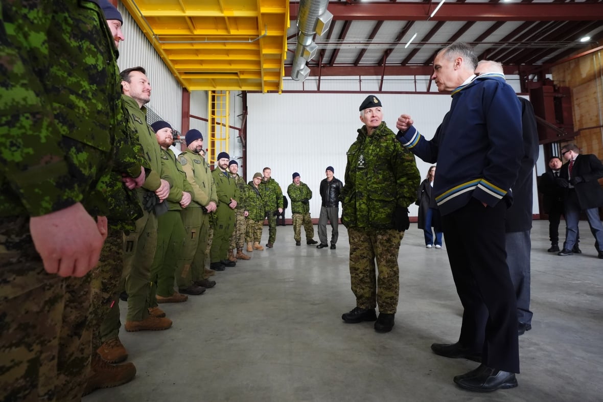 A man speaks to a line of military members indoors. An officer stands beside him.