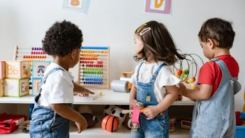 Getty Images Three young children have their backs turned to the camera as they stand beside a table full of toys. There are learning blocks, an abacus and a variety of sports equipment placed on top of or under the table. The children, two boys and a girl, are all wearing denim dungarees.