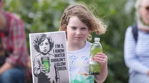 PA Media A young girl looks directly at the camera. In her right hand is a sign which reads 'I'm 5. I know water shouldn't glow green'. In her other hand is a plastic bottle which is full of green murky water.