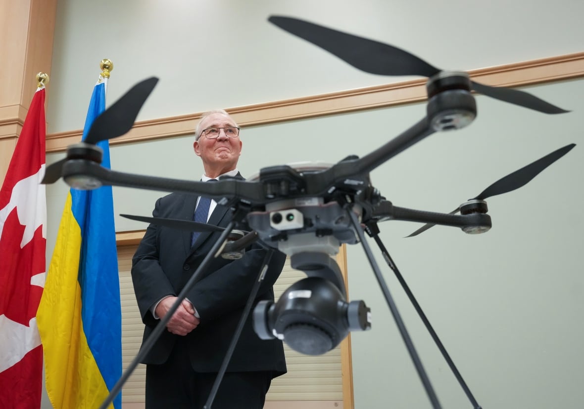 A man is seen standing beside a Canadian and Ukrainian flag. A drone is seen in front of him.