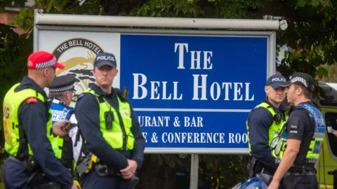 EPA/Shutterstock Police officers wearing yellow hi-vis jackets and caps standing in front of a large blue and white sign that says "The Bell Hotel".