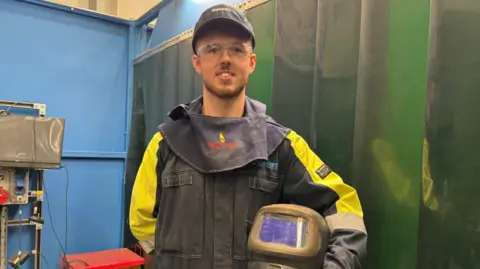 Laurence in welding protective clothing wearing a cap, black and yellow overalls and goggles, holding a helmet under his arm. He is looking at the camera.