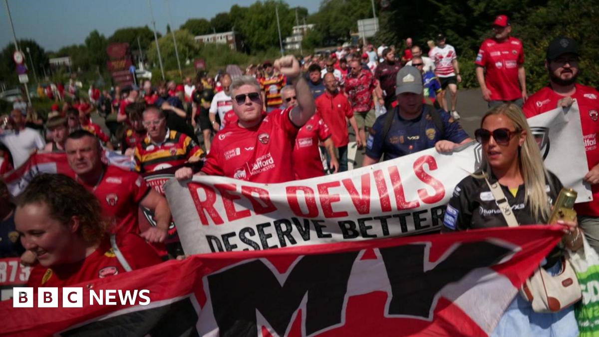A crowd of Salford Red Devils fans march together, dressed in the club's red and black colours. One banner reads "Red Devils deserve better".