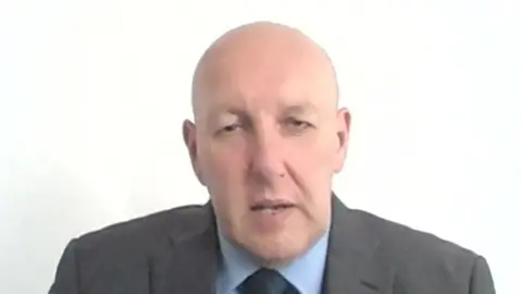 Mark Fairhurst, who is bald, clean shaven and wears a grey suit over a blue shirt, speaks into a laptop camera against a white background. 