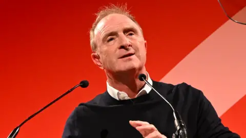 Edinburgh TV Festival James Harding speaking at the Edinburgh TV Festival. He has short red hair and wears a white shirt under a thin black jumper. He has a neutral expression as he speaks into two small black microphones in front of a plain, bright red background.