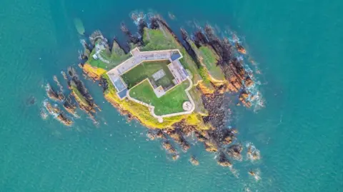 Strutt & Parker An aerial view looking directly down on to the island. The stone outer walls surround the majority of the land, with a green courtyard inside. Cliffs and rocks surround it, the sea is turquoise blue. 