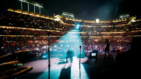 Lindsey Byrnes An image taken from the back of the stage as My Chemical Romance are performing. Gerard Way is in the centre, kneeling on the floor and facing the audience of thousands of people. He is under a spotlight, and his head is bowed forward. The stadium is lit up as it is nighttime.
