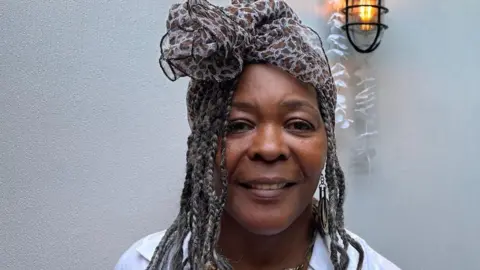 BBC Annetta Stanley is looking directly at the camera.  She has long, braided, hair and is wearing a scarf, tied in a bow, on her head.  She is wearing a white shirt and a gold coloured necklace. 