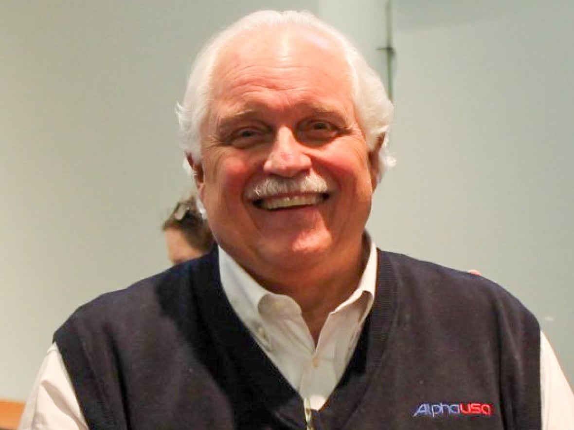 A man with short, white hair and a neat, white moustache smiles while wearing a sweater vest with the words AlphaPlus over a white collared shirt.