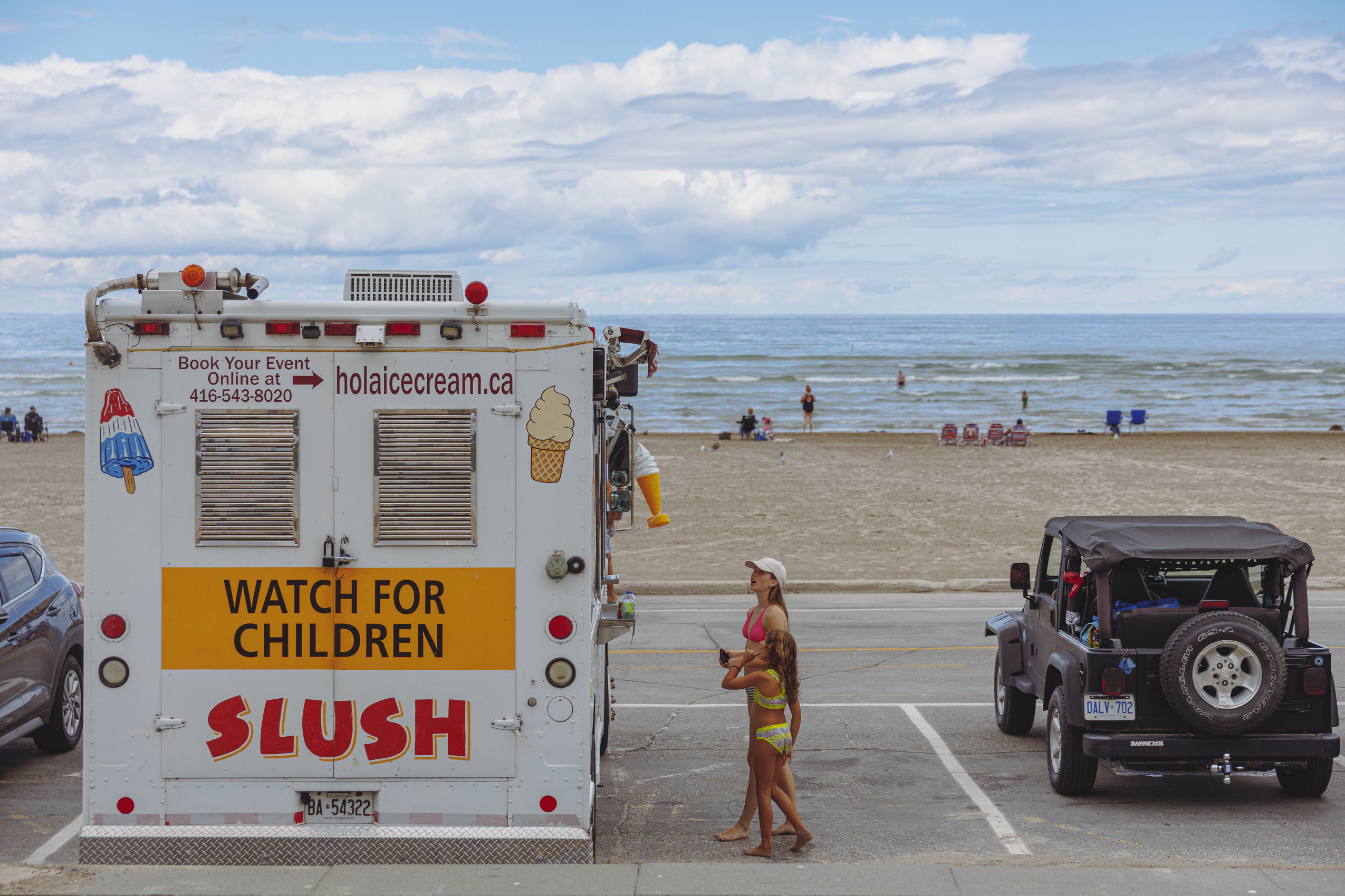 A woman and a girl in bathing suits approach an ice cream truck. In the background: Wasaga Beach, Lake Huron and a blue sky with clouds.