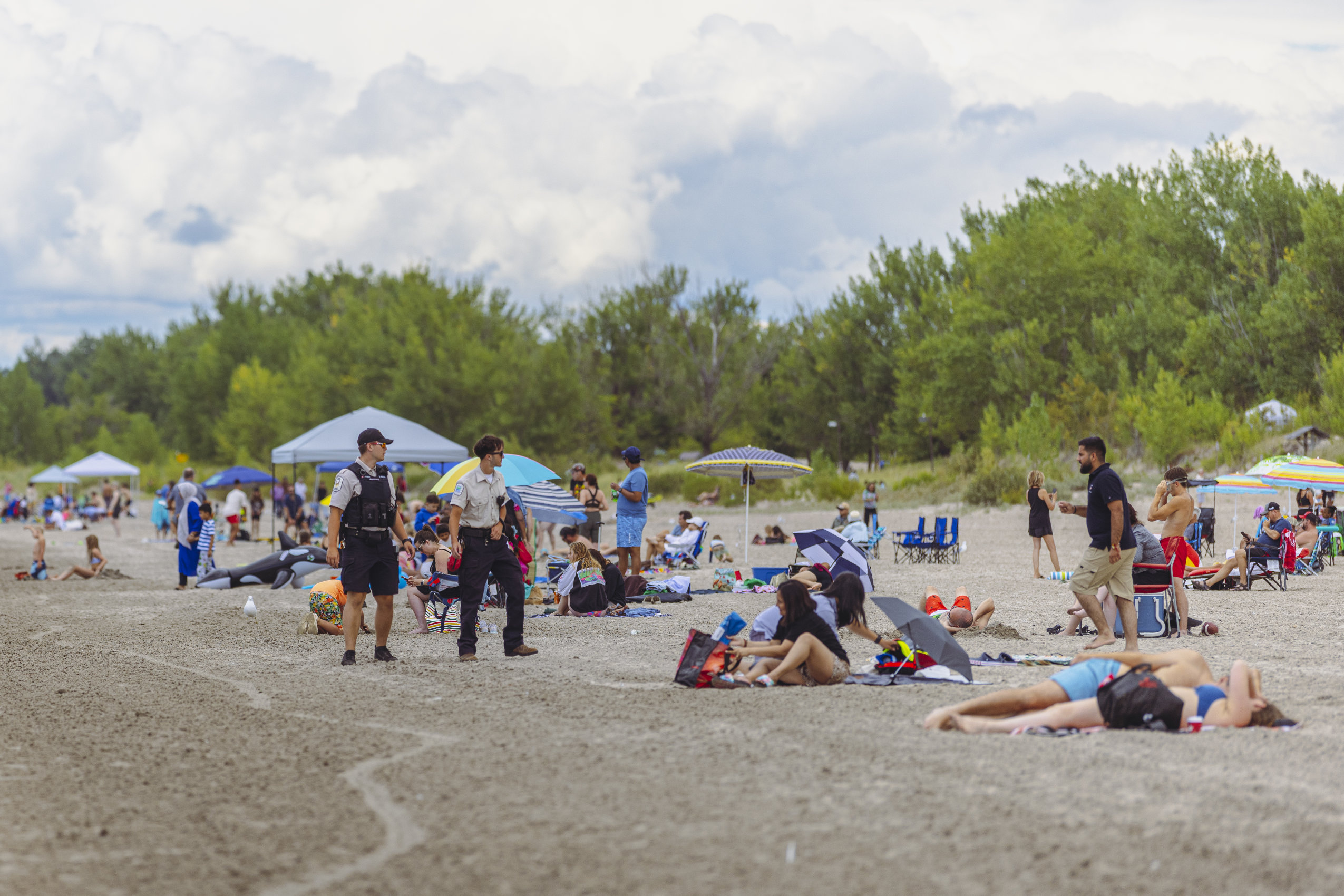 Two park wardens in official uniforms are seen walking along Wasaga Beach, as bathers loll about on the sand.
