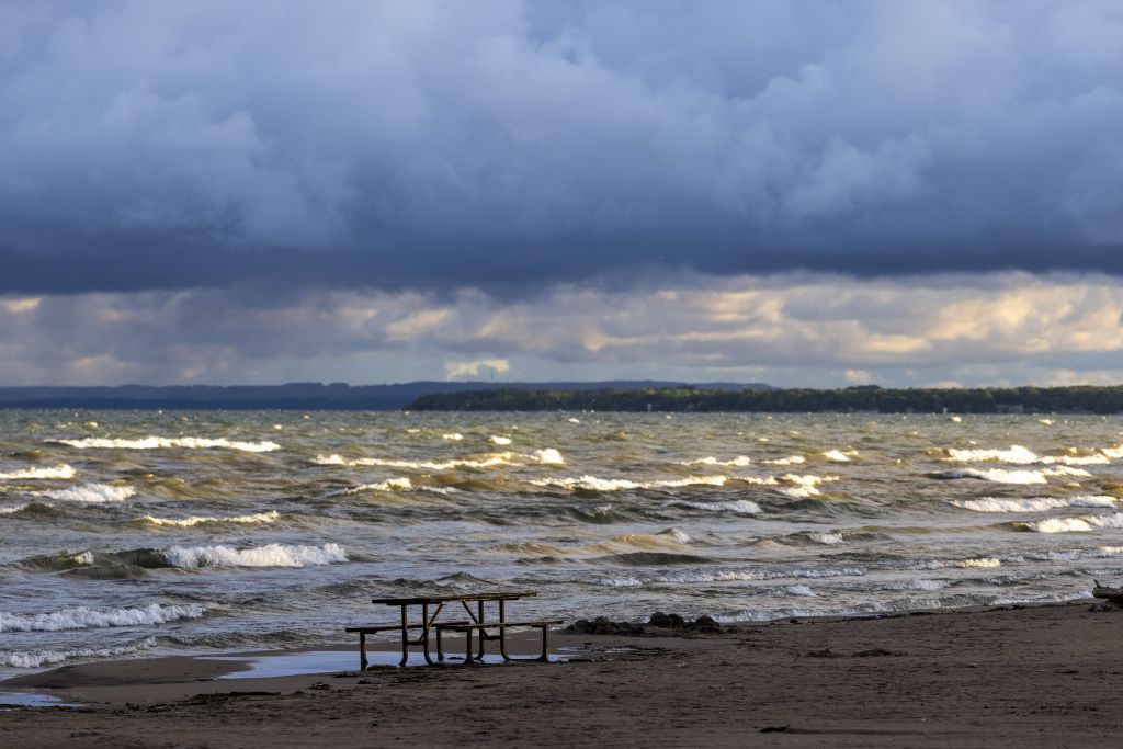 A cloudy sunrise drenches Lake Huron in dramatic lighting as whitecaps roll into the shore at Wasaga Beach. In the foreground, an empty picnic table sites on the beach.