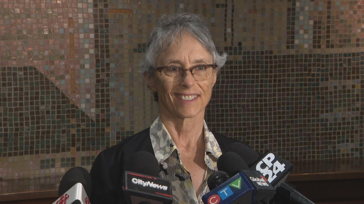 Woman in button-up shirt and black blazer stands in front of microphones, smiling