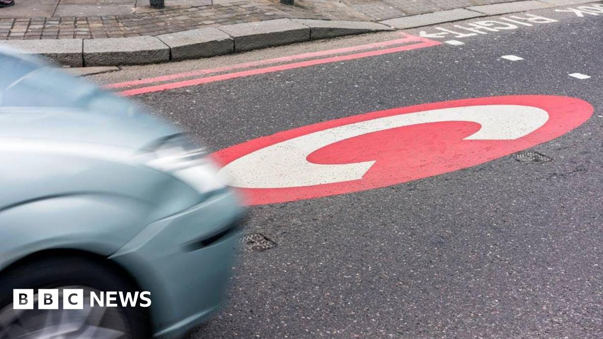 A red circle with a large white letter C is painted on the surface of a road in front of a crossing. A silver-blue car is driving towards the painted C from the left, and is blurred from motion. The pavement with double red lines along the kerb is visible in the background.