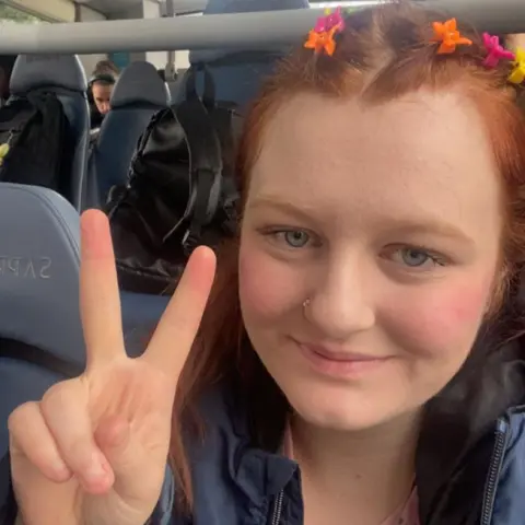 Maisy Moazzenkivi Maisy Moazzenkivi with short red hair with yellow orange and pink clips making a peace sign with her fingers while sitting on a bus