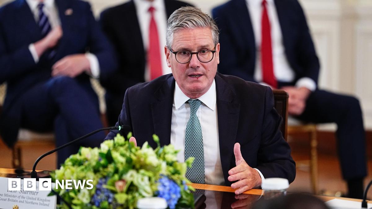 Keir Starmer sitting at glossed wooden table in White House meeting with other leaders. The camera is on him as he speaks. Starmer is gesturing with his left hand and is wearing a black blazer, a white shirt and a patterned blue tie. There are blue flowers in front of him on the table. Behind, sit three men against the wall - the heads are out of shot, but they are wearing suits, white shirts and a stripy navy and white tie, a red tie and a red tie respectively from left to right.
