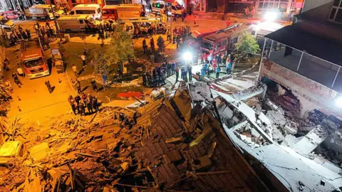 Berkan Cetin/Anadolu via Getty Images An aerial view of a collapsed building with emergency workers all around it. The picture is taken at night time and the debris is lit by strong spot lights.