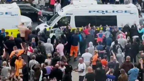 CCTV footage showing a crowd of people around a police van, several of whom have their hands against the sides of the vehicle, pushing it. Ross Hart is in the crowd, standing towards the front of the van wearing a brown T-shirt