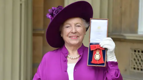 AFP via Getty Images Dame Stephanie Shirley wearing a purple hat, matching purple dress, and a pearl necklace, smiles at the camera while holding a medal outside Buckingham Palace after she was made a member of the Order of the Companions of Honour 
