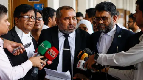 Reuters Prosecutor Tariqul Islam speaks to reporters after a hearing at a court in Dhaka. He is wearing a black suit and white shirt and surrounded by male journalists who hold microphones towards him.