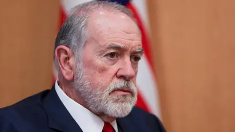 Reuters Mike Huckabee, who has grey hair and a short grey beard and moustache, looks on during an interview at his office at the US embassy in Jerusalem. An American flag is behind him.