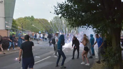 EPITOG A small crowd of protesters are turning away as a firework exploded in the car park of the Holiday Inn Express. The corner of the hotel building can be seen a few metres away.