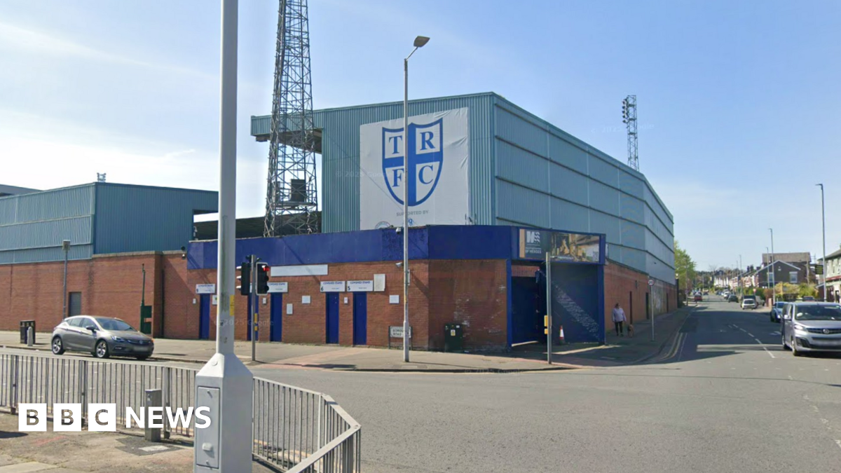 Google Streetview image of Tranmere Rovers' Prenton Park stadium. Taken from outside the ground, two large stands are visible. One of them includes a large blue-and-white shield with the initials TRFC printed on it.