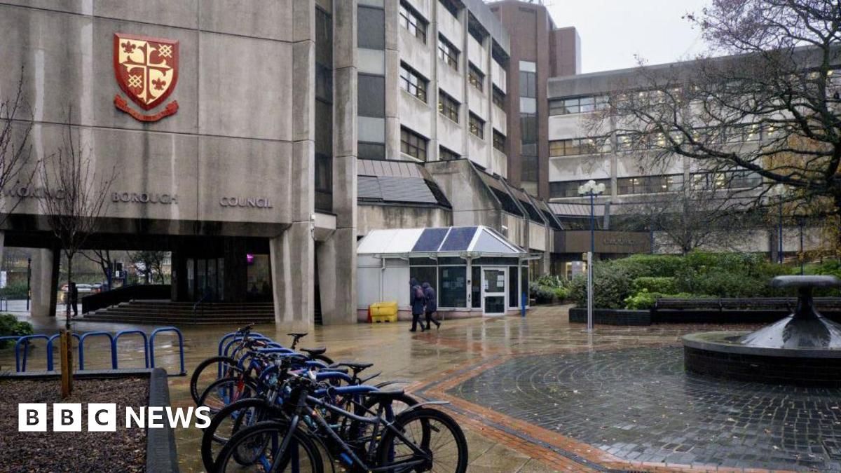 A grey, brutalist building on a rainy day. A red and gold Woking Borough Council logo is fixed to the side of the building and there are four bicycles locked to a railing outside the building.