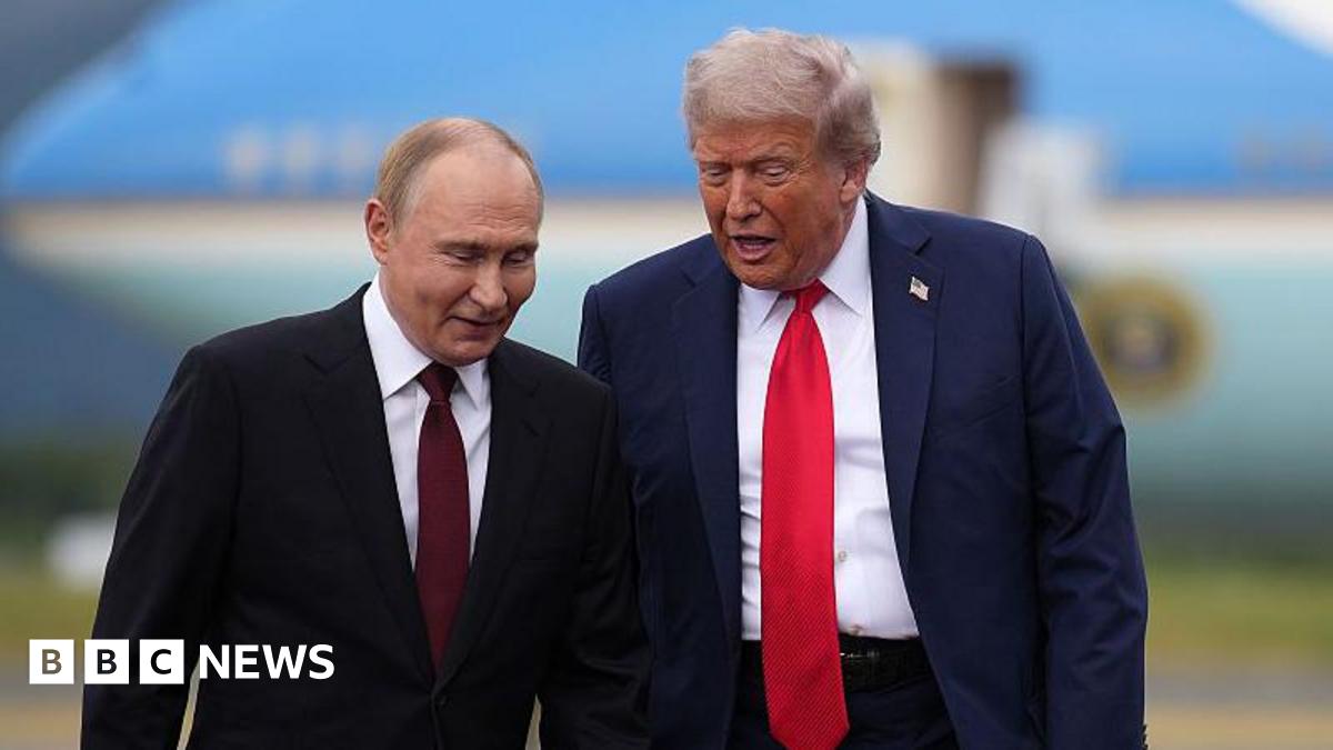 Vladimir Putin and Donald Trump pictured side-by-side on the tarmac of an airbase with a large blue aircraft blurred in the background. The men are mid-conversation and leaning in towards one another. Both are wearing dark suits with white shirts. Putin wears a maroon tie while Trump's is bright red. Trump also wears a pin of the US flag on his lapel.