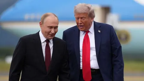 Getty Images Vladimir Putin and Donald Trump pictured side-by-side on the tarmac of an airbase with a large blue aircraft blurred in the background. The men are mid-conversation and leaning in towards one another. Both are wearing dark suits with white shirts. Putin wears a maroon tie while Trump's is bright red. Trump also wears a pin of the US flag on his lapel.