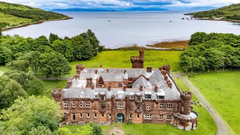Nest Marketing An aerial view of the red sandstone lodge with its ornate turrets and battlement-like rooftops. The lodge is set in an grounds with large areas of grass and trees. In the background is the sea, with a few boats dotted around a bay, and mountains in the distance.