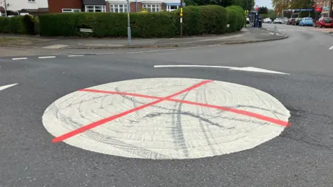 A large white mini roundabout circle in the middle of two roads. It has been graffitied with red spray paint to look like an England flag or St George flag