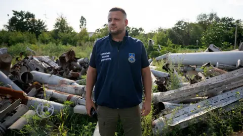 Lee Durant/BBC Dymtro Chubenko stands in front of a pile of Russian missile and drone parts