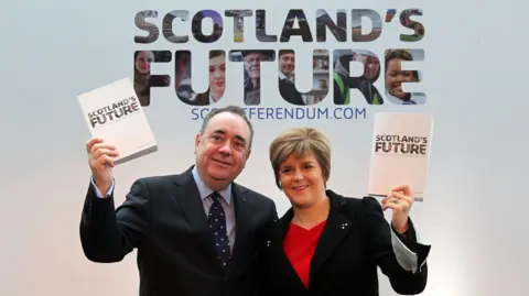 PA Media Alex Salmond on the left side wearing a dark suit over a blue shirt with a blue tie with a white pattern. He is holding a white book with the words 'Scotland's Future' on the front cover in dark writing. Nicola Sturgeon is on the right. She is wearing a dark jacket over a red top. She is also holding a copy of the book in her hand and raising it for the camera. The same words are behind them in a larger font in dark writing on a white background.