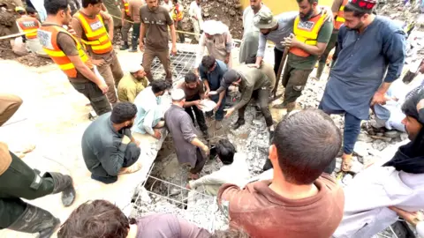 Rescuers clawing through rubble by hand. Men can be seen in high vis vests, and in more casual clothing. They are standing around a hole in the ground