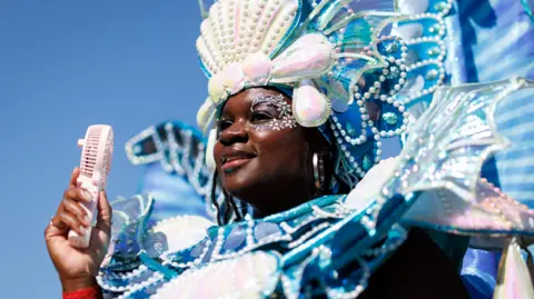 EPA A woman in a blue and white beaded costume holds a battery powered fan to her face.