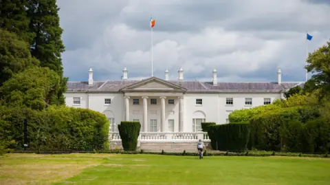 Getty Images An exterior shot of Áras an Uachtaráin. It shows a two-storey white building, resembling the White House in the USA. There is a flag pole with the Irish tricolour on it at full mast. Surrounding the building are large trees, bushes and a large patch of grass.