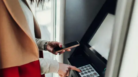Getty Images Woman getting cash out while holding her phone at a ATM