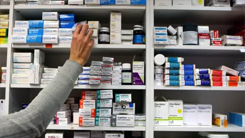 PA Media A person in a grey long-sleeve top reaching for a medication box on the top shelf in a pharmacy or medical storage area. The shelves are filled with neatly arranged boxes and containers of various medications, organised by type and brand.