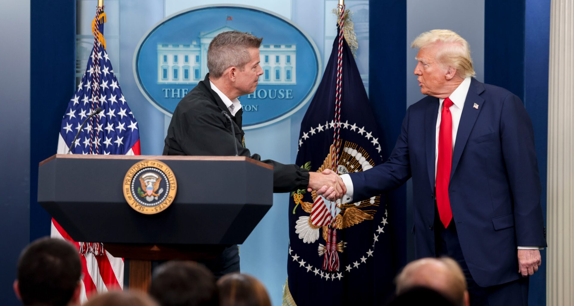 Two men wearing suits shake hands behind a podium