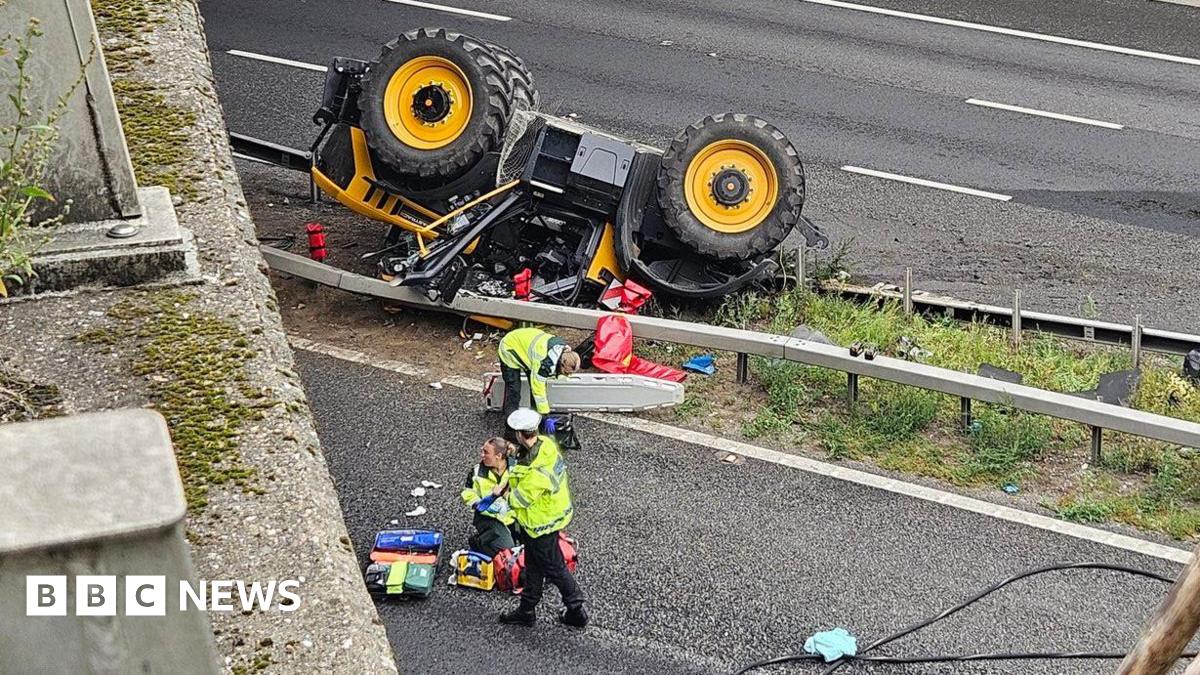 A yellow tractor on its roof under a bridge. It is on the central reservation of a motorway.