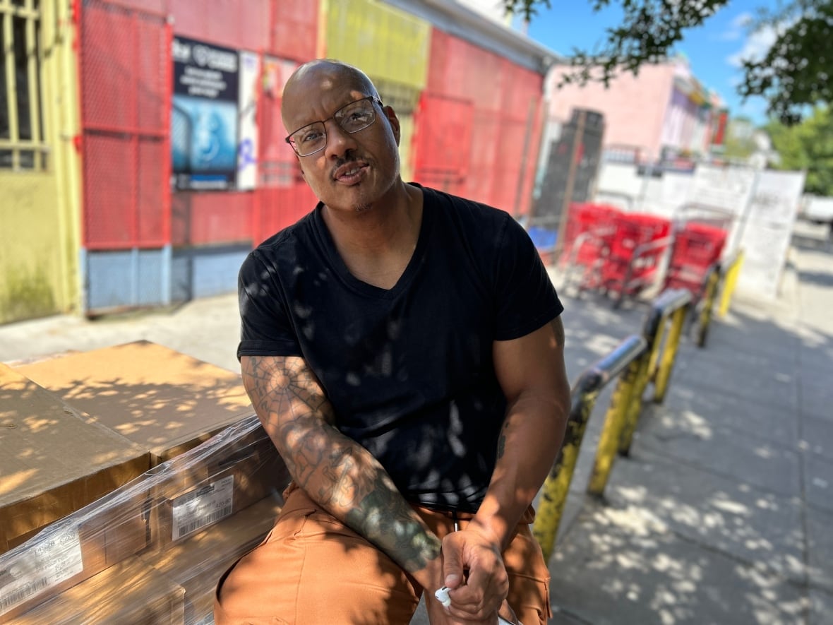 Damon is an African American man with a bald head. He's wearing rust coloured cargo pants and a back tshirt. He's sitting on a metal railing next to a crate of boxes. Behind him is a grocery store with red and yellow bars out front.
