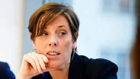 PA Media Jess Phillips looks on during a meeting as she wears a dark blue jacket. She has short, light brown hair and gold hooped earrings.