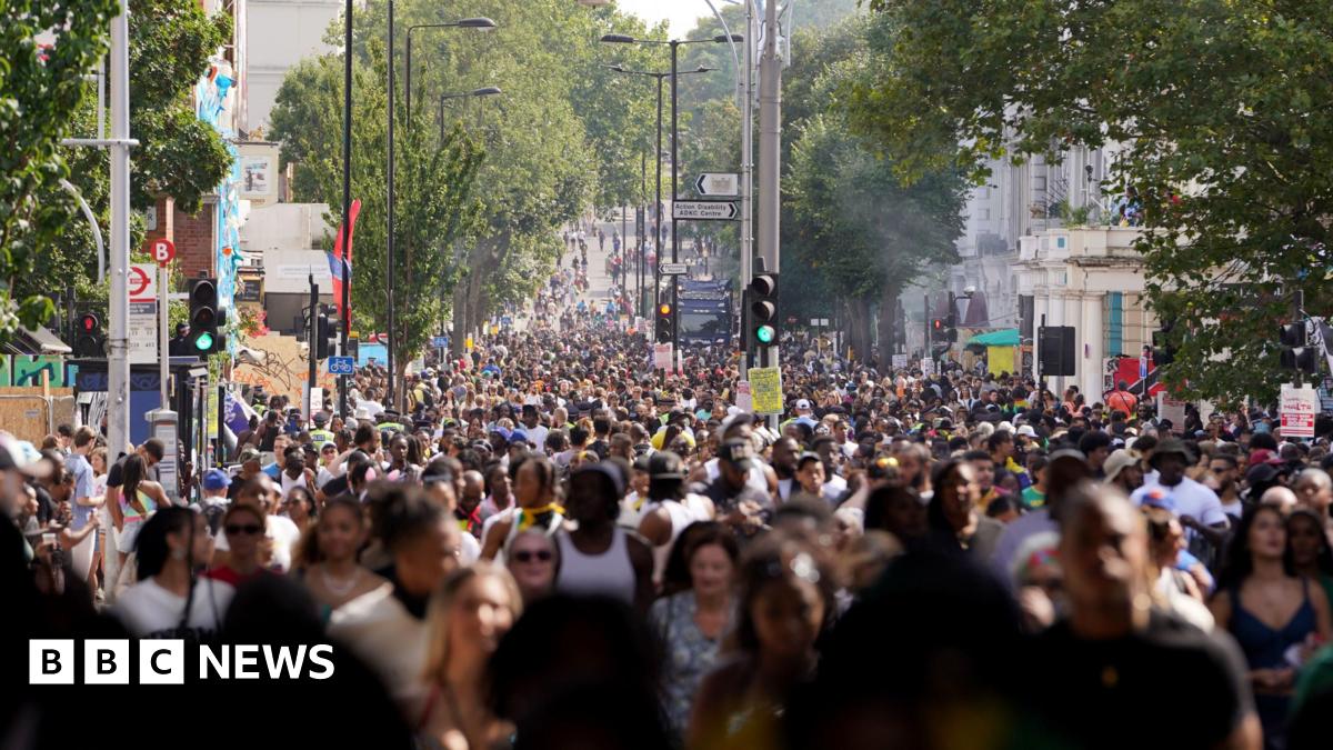 A sleek black drone with orange-tipped propellers sits on a white desk next to its remote control. The desk also has office items on it like telephones and cables.