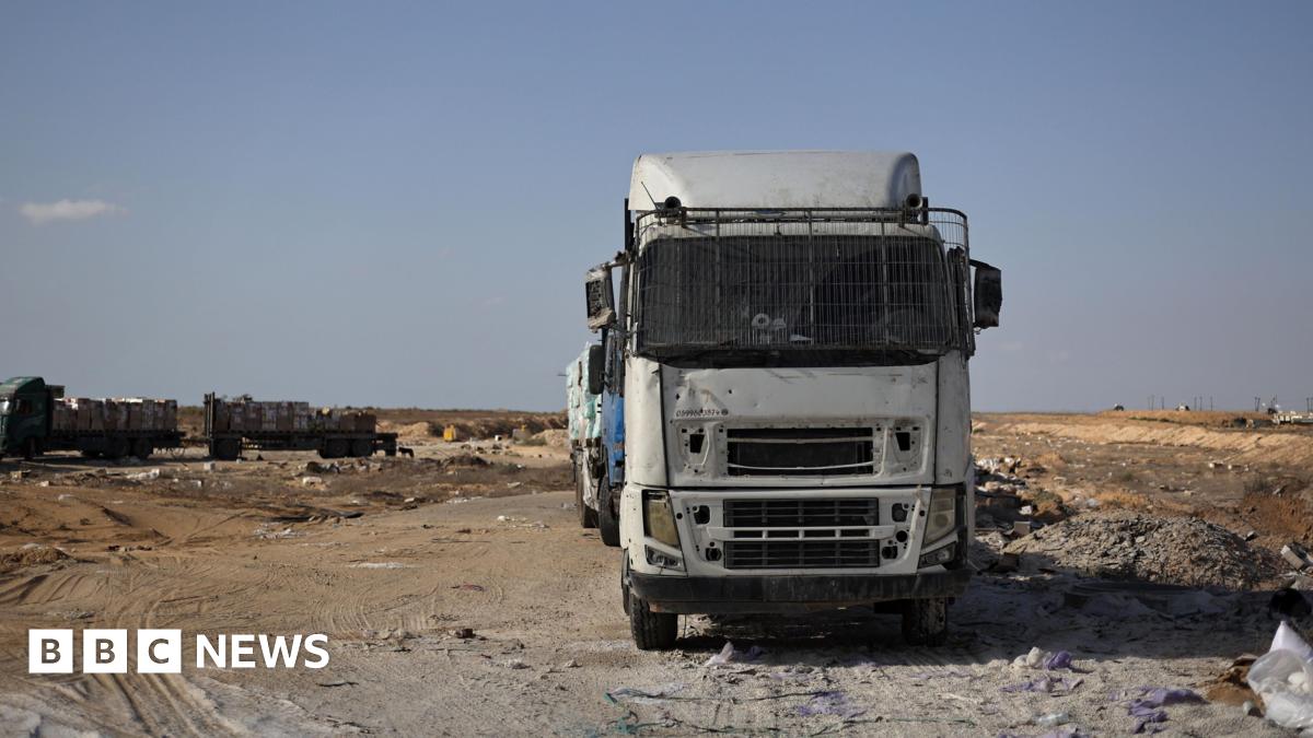 A white lorry waits on the Kerem Shalom border crossing in Gaza. More trucks can be seen in the distance. The foreground is a mix of arid ground and grey and brown sand.