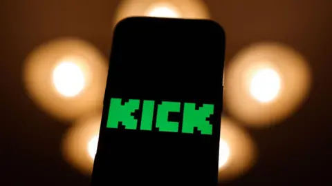 Getty Images A phone screen with the Kick logo in green on it. Black background with orange blurry lights. 